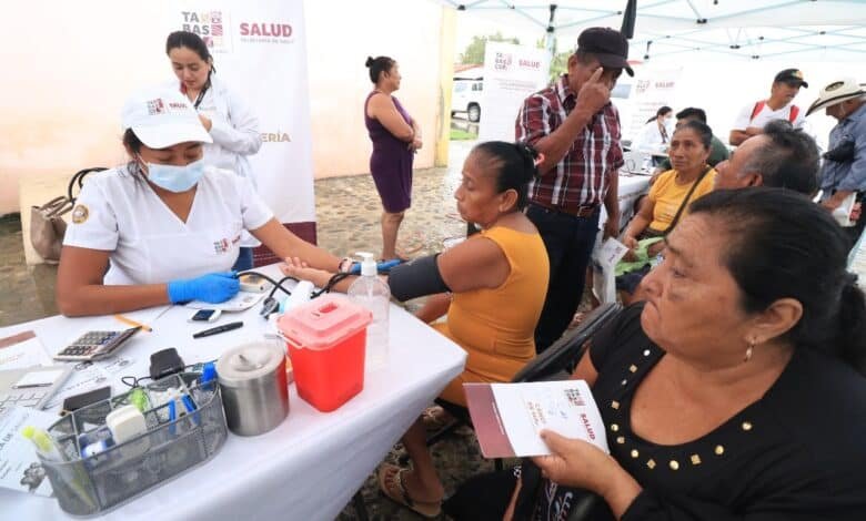 Persona recibiendo una toma de presión arterial en un módulo de salud, acompañada por personal médico con materiales informativos sobre la hipertensión. Cartel de fondo con el lema “Mida su presión arterial con precisión, contrólela, viva más”.